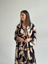 Vestido largo negro con estampado de hojas en tonos beige y tostado, de corte amplio y fluido, con escote en V y botonadura frontal parcial. Cuenta con mangas largas y volados en el faldón.
