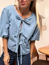 Blusa de lino color celeste con mangas abullonadas tres cuartos y cierre frontal mediante tres lazos decorativos tipo moño.