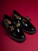 Mocasín de charol negro con plataforma, detalle de lazo y aplique de oso dorado.