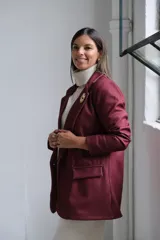 Blazer de corte sastrero en color bordó, con solapas clásicas, bolsillos frontales con solapa y cierre frontal con botones. Presenta una textura sutil en la tela y un broche decorativo en forma de sol en la solapa.