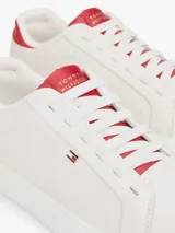 Zapatillas Tommy Hilfiger de diseño clásico y bajo, color blanco con detalles en rojo en la parte trasera del talón y en la lengüeta. Presentan cierre de cordones y el logo metálico distintivo de la marca en el lateral.
