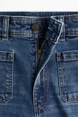 Jean de tiro alto con corte acampanado desde la rodilla. Presenta bolsillos tipo parche en la parte frontal y cierre con botón metálico.