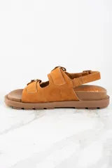 Sandalia de gamuza color tostado con base de goma marrón, diseño de tiras anchas con hebillas metálicas doradas y ajuste en el talón.