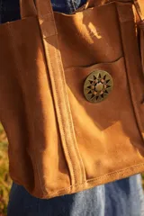 Cartera tipo tote de gamuza color marrón, con diseño amplio y relajado. Presenta un bolsillo frontal externo con cierre de broche metálico y una hebilla decorativa circular, además de un detalle de borla colgante en el lateral.