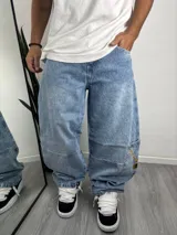 Pantalón de jean estilo baggy, color azul claro con efecto lavado. Presenta un diseño holgado, costuras reforzadas en las rodillas y un bolsillo lateral tipo cargo con un estampado gráfico de una cadena dorada y un candado con calavera.