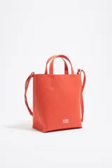 Bolso de mano de piel vacuna color naranja vibrante, con estructura blanda y tamaño mediano. Presenta dos asas superiores y una correa larga ajustable para llevar al hombro o cruzado. El exterior tiene textura granulada y el logo de la marca estampado en la parte inferior frontal.