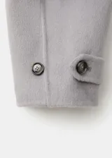 Chaqueta gris de diseño corto y holgado, con cuello alto envolvente, mangas anchas y puños ajustables con tiras y botones. Cuenta con bolsillos laterales y cierre frontal oculto.