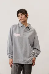 Buzo gris melange de cuello polo con cierre frontal de botones, mangas largas ranglán y aplique bordado rosado en el pecho.