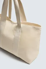Bolso tipo shopper de tejido de algodón color beige claro, con dos asas de mano y cierre magnético en el compartimento principal. Incluye un bolsillo interior con cierre de cremallera.