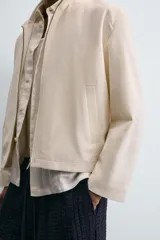 Cazadora de tejido color crema, con cuello alto, cierre frontal de cremallera y bolsillos de vivo en la cadera. Presenta un calce relajado (relaxed fit).