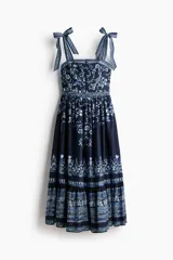 Vestido midi de algodón azul marino con estampado floral y geométrico en tonos celestes y blancos. Presenta escote recto, breteles ajustables con moñas, cuerpo entallado y falda acampanada con volados fruncidos en la parte inferior.