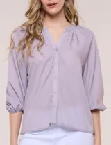 Blusa de gasa de color lila con calce holgado, cuello a la base con escote en V y cartera de botones a tono. Cuenta con fruncidos en el cuello y hombros que otorgan volumen, manga tres cuartos estilo abullonado con puño elastizado y ruedo suavemente redondeado.