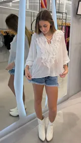 Top blanco de manga larga, confeccionado en tejido ligero y traslúcido. Presenta escote en V con cordones entrelazados y volados decorativos en el frente. Las mangas son largas y abullonadas con puños ajustados.