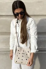Bandolera de cuero color beige con diseño de ojales metálicos distribuidos por toda la superficie frontal y placa metálica con logo de la marca. Cuenta con correa larga ajustable.
