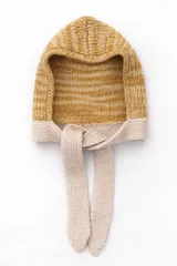 Capucha tejida a mano en lana merino, con diseño envolvente y lazos tejidos para anudar bajo el cuello. Presenta una combinación de colores en tonos mostaza y beige.