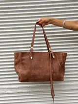 Cartera tipo tote de cuero color marrón, con diseño de doble asa larga y detalles de hebillas metálicas plateadas. Incluye flecos decorativos laterales.