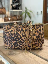 Cartera tipo tote con estampado animal print de leopardo. Cuenta con doble asa de hombro ajustable con hebillas metálicas y detalle de flecos colgantes en uno de los laterales.