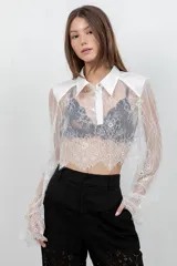 Camisa crop top de encaje blanco con mangas largas acampanadas, cuello camisero y pechera de tela lisa.