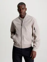 Campera tipo bomber de Calvin Klein en color beige, confeccionada en una mezcla de algodón y poliamida. Presenta cuello alzado, puños y bajo de punto canalé, cierre frontal de cremallera, bolsillos laterales de hendidura y un bolsillo adicional con cremallera en la manga izquierda con insignia de goma de la marca.