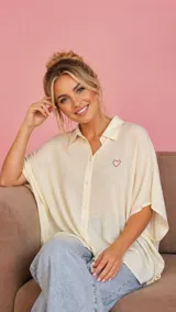 Camisa tipo vestido de corte holgado, confeccionada en viscosa suave, con cuello camisero, cierre frontal de botones, mangas cortas con dobladillo y bolsillos laterales. Presenta un pequeño bordado de corazón en el pecho.