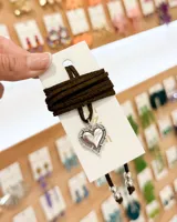 Choker de tiento de gamuza color marrón oscuro con dije de corazón plateado con borde trabajado. El diseño incluye terminaciones con cuentas metálicas plateadas en los extremos.