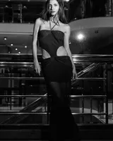 Vestido largo negro, ajustado al cuerpo, con escote strapless y tirantes finos cruzados en el cuello. Presenta recortes laterales pronunciados en la cintura y un nudo fruncido en la parte inferior frontal.