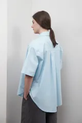 Camisa de corte oversize y diseño unisex, color celeste, con cuello clásico, manga corta amplia y bolsillo frontal. En la espalda presenta un canesú con pliegue central que aporta amplitud y caída fluida.