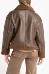 Campera tipo gamulán de cuero sintético en color marrón, con interior y detalles en cuello y puños de corderito sintético al tono. Presenta un corte oversize con cierre frontal metálico.