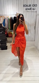 Vestido largo rojo con diseño asimétrico, escote redondo y detalle de nudo drapeado en la cintura que crea una abertura frontal, dejando ver una falda corta interior.