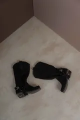 Bota estilo tejana de caña alta, confeccionada en cuero marrón con acabado texturizado. Presenta punta cuadrada, taco bajo y una correa con hebilla metálica ajustable y estribo desmontable en la zona del tobillo.