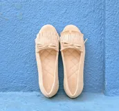 Mocasín de cuero gamuzado color avena, con detalle de flecos en la capellada y un lazo frontal con herrajes metálicos dorados. Cuenta con interior forrado en cuero y suela de goma flexible.