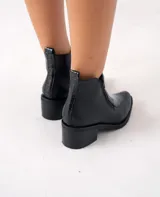 Bota corta de cuero vacuno color negro, con diseño combinado de cuero liso y cuero con textura de reptil en el empeine. Presenta punta fina, taco cuadrado de 6 cm y doble cierre lateral.