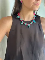 Collar corto con cuentas de nácar teñido en fucsia y turquesa, piedras naturales en verde, cuentas de acrílico en fucsia y mostacillas rojas. Se cierra con cadena de acero quirúrgico.