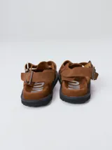 Slipper tipo guillermina confeccionada en gamuza color marrón, con plataforma de goma negra y doble hebilla ajustable. Presenta un pespunte decorativo en el borde inferior y cuenta con interior recubierto en cuero.