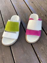 Sandalias bajas de cuero blanco con dos tiras: una tira ancha de gamuza verde en una sandalia y rosa en la otra, y una tira fina de cuero blanco.