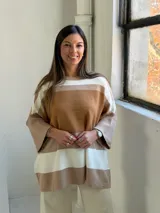 Poncho de punto con diseño de rayas horizontales en tonos tierra, blanco y beige. Presenta un corte holgado, cuello redondo y mangas anchas con puños acanalados.