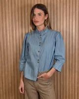 Camisa de denim celeste con rayas verticales blancas, cuello alto con volados, mangas tres cuartos abullonadas en los hombros y cierre frontal con botones oscuros.