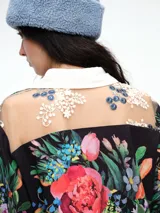 Camisa de corte relajado con estampado floral vibrante sobre fondo negro. Presenta cuello clásico, cierre frontal con botones y puños en contraste. El diseño destaca por sus terminaciones redondeadas y delicadas.