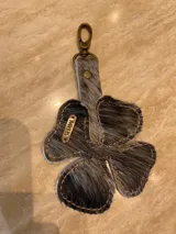 Llavero en forma de trébol de cuatro hojas confeccionado en cuero vacuno con pelo, en tonos grisáceos y negros. Incluye un mosquetón metálico en color bronce y una placa metálica pequeña con la marca grabada.
