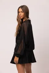 Campera bomber negra con transparencias, confeccionada en gasa con seda labrada. Presenta cierre frontal metálico, bolsillos cargo frontales con tachas y tiras colgantes, mangas con volumen y puños y cuello de rib elástico.