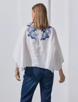 Blusa blanca con bordado floral azul, escote en V, manga japonesa y cierre frontal con botones.