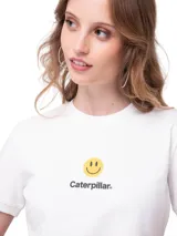 Remera blanca de corte crop con cuello redondo y mangas cortas. Presenta un estampado frontal con el logo de Caterpillar y un ícono de cara sonriente en color amarillo. Incluye una pequeña etiqueta negra con el logo de la marca en el dobladillo inferior.