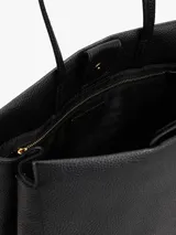 Cartera tipo tote de Tommy Hilfiger en color negro, confeccionada en poliuretano con textura granulada. Presenta doble asa superior, cierre principal y un diseño central decorativo con tachas metálicas y el logo monograma TH en metal dorado.