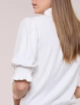 Buzo de algodón blanco con cuello mao y volados, manga tres cuartos abullonada y botones metálicos decorativos en la parte frontal.