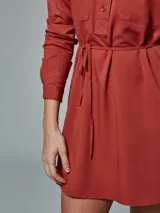 Vestido camisero color verde óxido, con cuello camisero, mangas largas con puños abotonados, dos bolsillos con solapa en el pecho, cinturón de lazo y cierre frontal con botones.