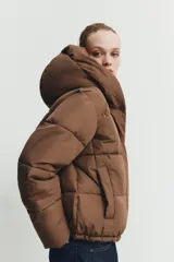 Campera puffer acolchada de color negro, con cuello envolvente, capucha integrada, manga larga, bolsillos laterales ocultos y bajo con elástico. Cierre frontal con botones a presión ocultos por solapa.