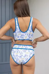 Conjunto de bikini blanco con estampado floral en tonos azules y amarillos, con ribetes azules. El corpiño es de triángulo con breteles regulables y la bombacha es de tiro alto con doble banda elástica.