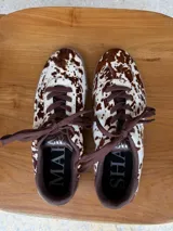 Championes urbanos con diseño de estampado animal print tipo vaca, confeccionados en cuero con textura de pelo. Presentan cordones marrones, suela de goma en tono tostado y detalles en color marrón oscuro en el cuello y lengüeta.