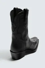Bota estilo cowboy de caña media, confeccionada en piel negra con costuras decorativas en la caña y el empeine. Presenta una horma en punta, tacón ancho y tiradores laterales.