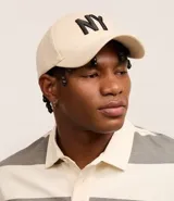 Gorra de seis paneles con ala curva, confeccionada en algodón color marrón. Presenta un bordado frontal con las letras NY en color blanco y cuenta con cierre strapback ajustable en la parte posterior.